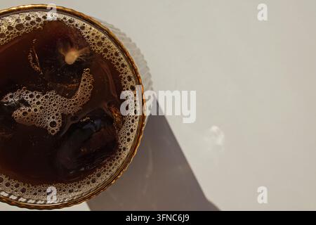 Vista dall'alto di caffè freddo nero con schiuma in un bicchiere rigato con bordo dorato che proietta un'ombra diagonale dura, spazio per il testo. Foto Stock