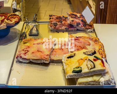 Fette assortite di pizza italiana ricoperte di formaggio, prosciutto, olive e verdure grigliate esposte su un vassoio di metallo in una pizzeria locale. Foto Stock
