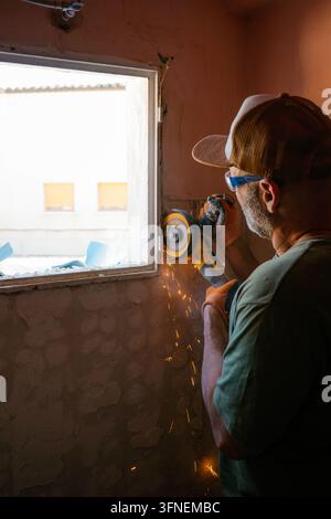 Uomo di mezza età con occhiali protettivi e guanti, che taglia una cornice di finestra con una smerigliatrice angolare Foto Stock