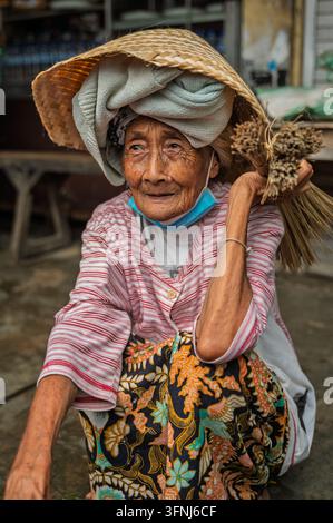Ritratto di una donna indonesiana anziana con un cappello intrecciato e abiti a fantasia, che porta una scopa fatta a mano sulla spalla in un mercato tradizionale. Foto Stock