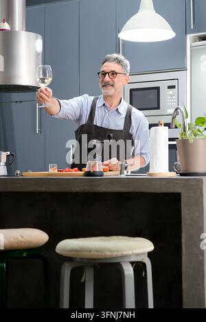Uomo maturo con grembiule prepara un pasto a casa mentre solleva un bicchiere di vino bianco in una cucina moderna e stilosa con elettrodomestici, verdure e c Foto Stock
