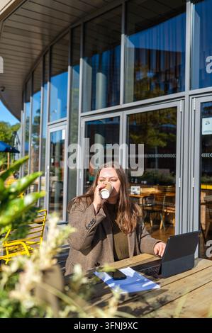 Freelancer donna beve caffè mentre lavora su un laptop a un tavolo di un caffè all'aperto, circondata da documenti e uno smartphone accanto a un moderno edificio in vetro Foto Stock