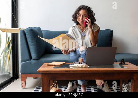 Donna con vitiligo seduta su un divano a casa, esamina documenti mentre parla al cellulare accanto a un laptop aperto, illustra il lavoro da remoto e la sera Foto Stock
