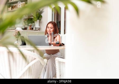 Giovane donna usa un laptop seduta a un tavolo in un interno domestico luminoso, incorniciata da un primo piano foglioso morbido e circondata da piante d'appartamento e natura Foto Stock
