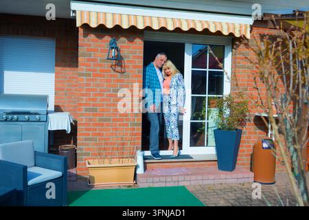 Coppia felice di mezza età in piedi insieme sulla porta della loro casa di mattoni, godendosi un momento rilassato sul patio accanto ai mobili da esterno, grill acceso Foto Stock
