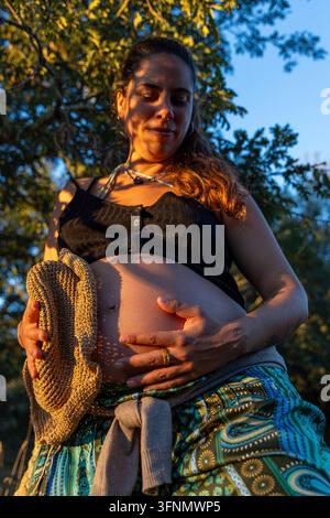 Donna ispanica incinta in piedi all'aperto, tocca delicatamente il ventre mentre si crogiola nella luce dorata del sole dell'ora dorata Foto Stock