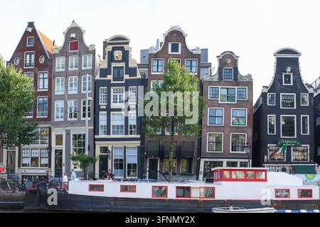 Amsterdam, Paesi Bassi - 1 ottobre 2026: Case strette tradizionali sul canale e una casa galleggiante ormeggiata costeggiano il lungofiume storico. Foto Stock