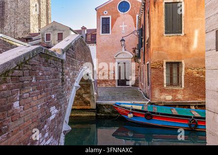 Barca tradizionale in legno colorata ormeggiata vicino a un ponte di mattoni e una piccola facciata di chiesa lungo un canale tranquillo a Venezia, regione del Veneto, Nord Italia Foto Stock