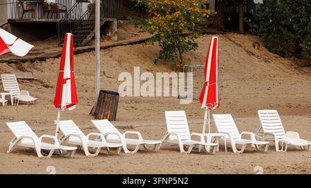 Una vista esterna di una spiaggia di resort sul lago vuota con file di sdraio in plastica bianca e ombrelloni rossi chiusi sulla sabbia, con un logoro Foto Stock