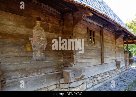 Esterno di una chiesa in legno storica che mostra un'antica pittura di un santo religioso su un muro di tronchi sopra un vicolo fondato su pietre di fiume Foto Stock