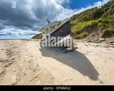 Relitto di una piccola barca da pesca bianca e blu arenata sul sabbia della spiaggia accanto a una duna alla spiaggia di Cambois, Northumberland Foto Stock