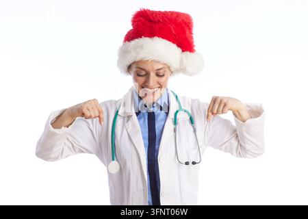 Una giovane dottoressa con gli occhi chiusi e un sorriso gioioso indossa un cappello di Babbo Natale e una camicetta da laboratorio, indicando le sue spalle in un gesto festivo delle festività Foto Stock