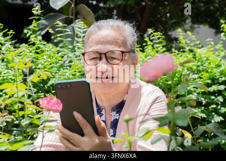 Donna asiatica anziana nel suo giardino con occhiali e che usa lo smartphone, forse per scattare foto o fare una videochiamata. Foto Stock