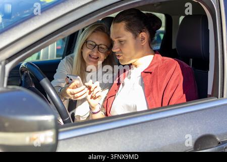 Donna bionda sorridente con occhiali si china verso un uomo in camicia rossa seduto al volante mentre guardano insieme uno smartphone. Concetto di piano Foto Stock