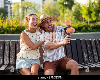 Coppia giovane che ride e posa per una fotocamera smartphone mentre si gode il tempo all'aperto. Foto Stock
