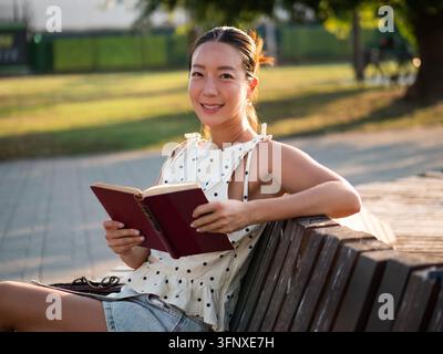Donna asiatica di mezza età felice che si rilassa nel parco, legge un libro, guarda in camera e sorride. Foto Stock