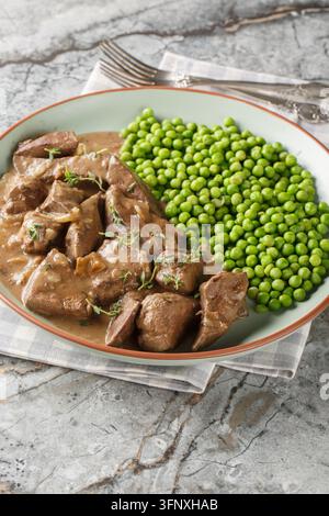 Fegato di manzo alla badenna tedesca con cipolle, stufato nel vino bianco e servito con piselli verdi, primo piano su un piatto su un tavolo. Verticale Foto Stock