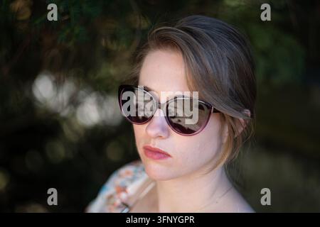 Ritratto ravvicinato di una giovane donna che indossa occhiali da sole scuri e alla moda, che guarda lontano all'aperto con uno sfondo sfocato naturale e delicato alla luce del giorno Foto Stock