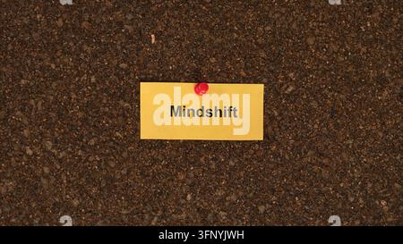 Un biglietto di carta giallo con la parola Mindshift stampata sopra è appuntato con una graffetta rossa su uno sfondo di pannello di sughero marrone. Foto Stock