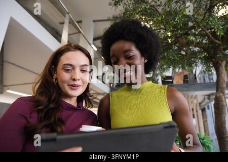 Due colleghe donne in piedi nell'atrio dell'ufficio con magliette marrone e verde, tengono un tablet e un caffè Foto Stock