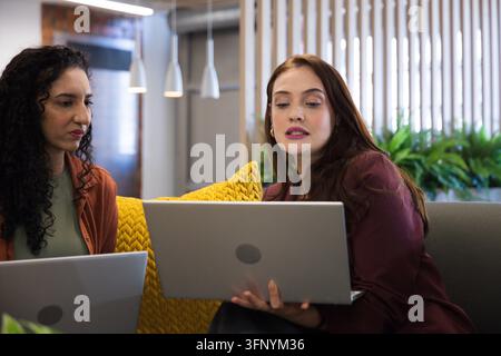 Diverse colleghe donne che presentano su due laptop mentre sono sedute su un divano grigio in blazer Foto Stock