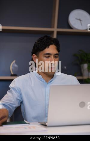 Uomo asiatico che lavora su un laptop argentato alla scrivania dell'ufficio, indossa una camicia blu e smista carte Foto Stock