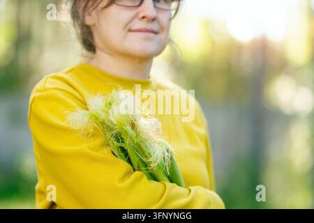 Una donna in maglione giallo guarda la telecamera con un fiore tra i capelli in un ritratto ravvicinato. La modella serena che posa con un'espressione gentile presenta una visione elegante nella luce naturale soffusa. Foto Stock