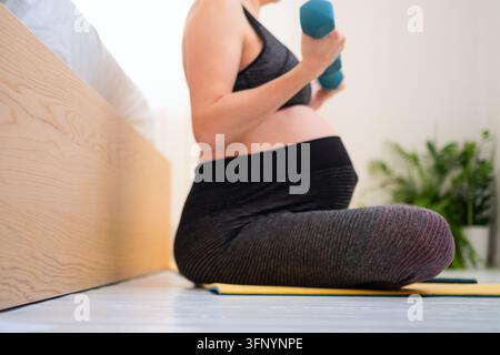 Una donna incinta è seduta su un tappeto da yoga tenendo un manubrio in un ambiente calmo e tranquillo a casa. La madre che pratica yoga mostra una serena consapevolezza e un rilassamento equilibrato e sano al chiuso. Foto Stock