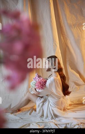 la donna è seduta sul pavimento, tiene un mazzo di fiori. Ha i capelli lunghi e indossa un abito fluido. Una luce soffusa illumina la scena, crea Foto Stock