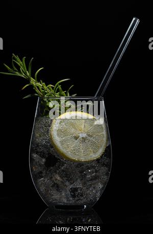 Cocktail Gin Tonic con ghiaccio, fetta di limone, rametto di rosmarino fresco e cannuccia di vetro in un bicchiere tumbler su sfondo nero Foto Stock
