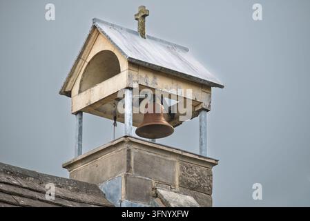 Campanile con una campana di bronzo e una croce coperta di muschio sul tetto metallico. La struttura in legno con vernice scrostata poggia su una base in pietra Foto Stock