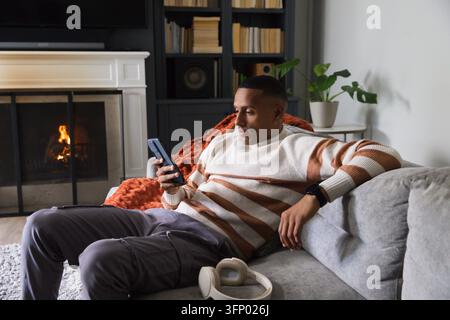 Uomo afroamericano sdraiato su un divano grigio accanto al caminetto, indossa un maglione a righe e tiene in mano uno smartphone Foto Stock