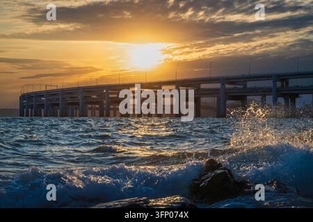 Le onde si infrangono contro gli scogli costieri sotto le campate di avvicinamento del ponte della baia di Xinghai al tramonto a Dalian, Liaoning, Cina. Foto Stock