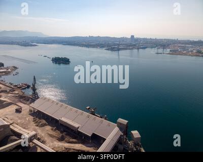 Il porto industriale si trova accanto a una città costiera estesa, con gru di carico, cumuli di aggregati e lunghi capannoni di stoccaggio coperti lungo l'acqua. Piccola foresta è Foto Stock
