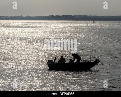 Due uomini adulti si godono una giornata di pesca da un piccolo motoscafo su uno specchio d'acqua scintillante, con il sole che si riflette sulla superficie e una massa terrestre lontana visibile sotto un cielo soffice Foto Stock