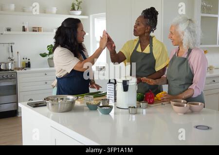 Diverse amiche donne che si danno il cinque mentre preparano un pasto in cucina indossando grembiuli con un fornello elettrico Foto Stock