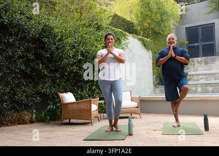 Coppia di anziani che pratica yoga in piedi su materassini verdi in terrazza indossando abiti sportivi con bottiglie d'acqua Foto Stock