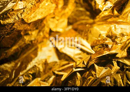 La carta d'oro accartocciata riempie l'inquadratura, creando pieghe angolari, riflessi luminosi, ombre profonde e una trama metallica astratta senza oggetti visibili Foto Stock