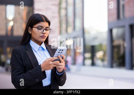 Giovane donna d'affari indiana in abito e occhiali, in piedi all'aperto in un contesto urbano, concentrata sul suo telefono cellulare e attivamente messaggiando o navigando Foto Stock