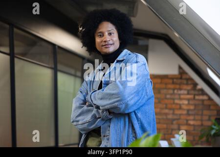Uomo afroamericano in piedi con le braccia incrociate in ufficio vicino a una pianta in vaso, indossa una giacca di jeans Foto Stock