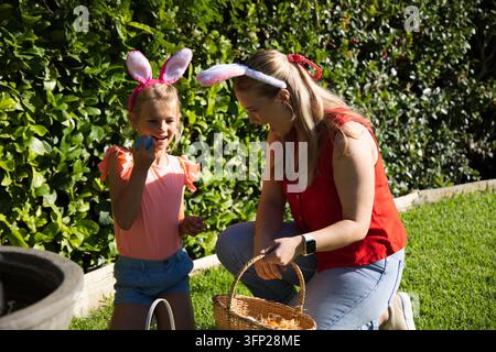 Mamma e figlia con orecchie da coniglio che festeggiano in giardino tenendo un cesto intrecciato con uova Foto Stock