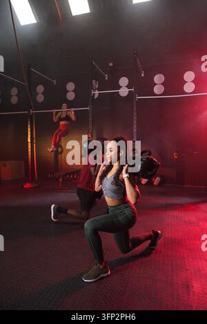 Gruppo di palestra diversificato che esegue affondi con pesi mentre una donna in top corto grigio e leggings verdi tiene un sacco di sabbia Foto Stock
