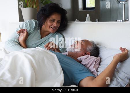 Coppia di anziani sorridente mentre si appoggia su una testiera imbottita nella camera da letto con copriletto bianco, un anello visibile Foto Stock