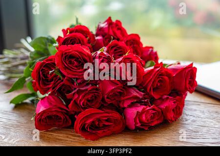 Mazzo di rose rosse fresche disposto su un tavolo di legno vicino alla finestra, composizione floreale romantica con petali vivaci, foglie verdi e luce naturale soffusa. Foto Stock