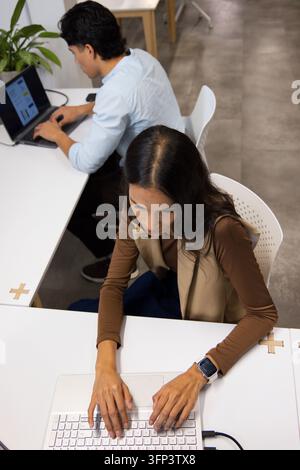 Diversi colleghi che digitano su laptop a scrivanie da ufficio moderne, donna che indossa un gilet beige Foto Stock