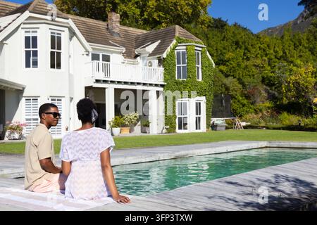 Coppia afroamericana seduta e in conversazione in piscina su un ponte di legno con asciugamano per occhiali da sole Foto Stock