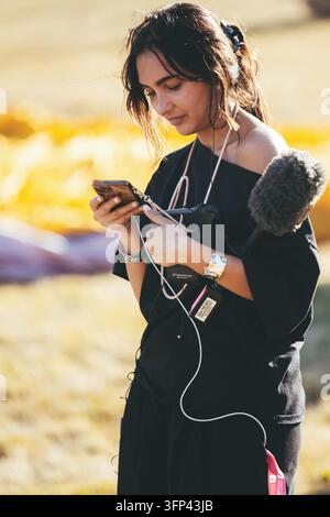 Bristol Balloon Fiesta, 7 agosto 2026, UK - Reporter televisiva di ITV News che controlla uno smartphone mentre tiene un microfono su un campo festivaliero soleggiato Foto Stock