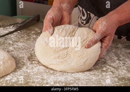Le mani di un panettiere lavorano attivamente su una porzione di impasto crudo ricoperta di polvere di farina fine su un piano di lavoro macchiato, pronta per il passo successivo durante la cottura Foto Stock