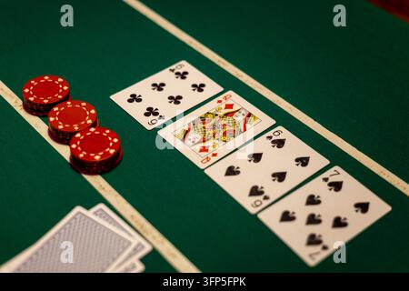 Tavolo da poker verde con quattro carte aperte allineate accanto a pile di gettoni rossi. Il tabellone mostra sei di fiori, donna di quadri, sei di picche. Foto Stock