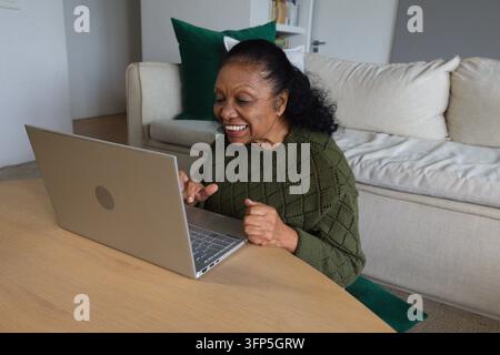 Donna anziana africana appoggiata, che digita su un laptop argentato a un tavolo nel soggiorno, indossa un maglione verde Foto Stock